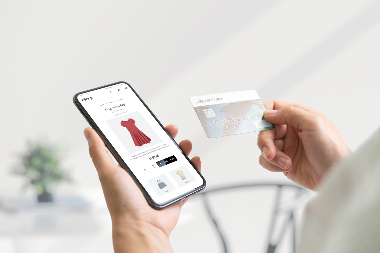 Ein EGIDO-Mandant hält ein Smartphone in der Hand, auf dem ein rotes Kleidungsstück angezeigt wird. In der anderen Hand befindet sich eine Kreditkarte, während der Kaufprozess auf einer E-Commerce-Webseite fortgesetzt wird.